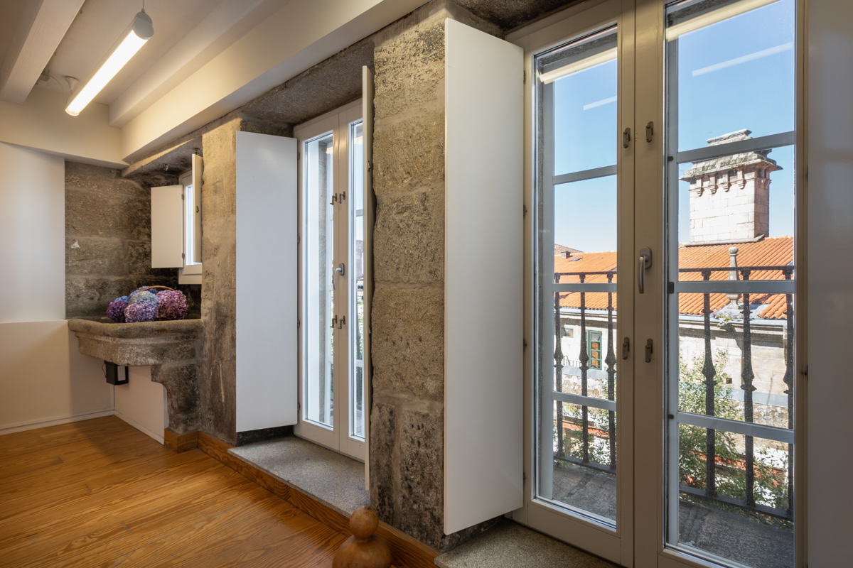 Fotografía del interior de una habitación con muros de piedra y un antiguo lavadero, y dos balcones con vistas a la calle.