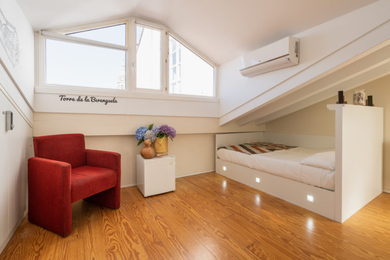 Fotografía del interior de la habitación del ático con cama individual con iluminación personalizada, un sillón, una nevera, aire acondicionado, taquilla con seguro y ventanas..