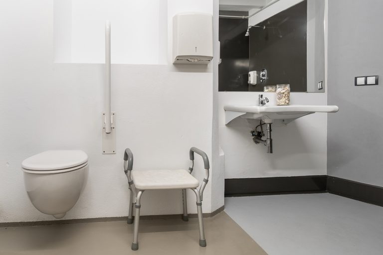 Fotografía de baño adaptado para personas que usen silla de ruedas, con váter, silla, lavabo, espejo, y reflejado en el espejo se aprecia la ducha.