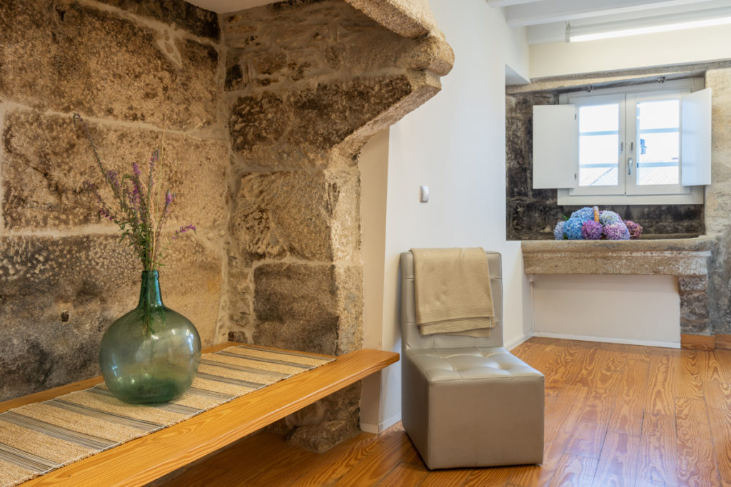 Fotografía del interior de una habitación con muro de piedra y un lavadero antiguo, una silla junto a una repisa con un jarrón decorativo.