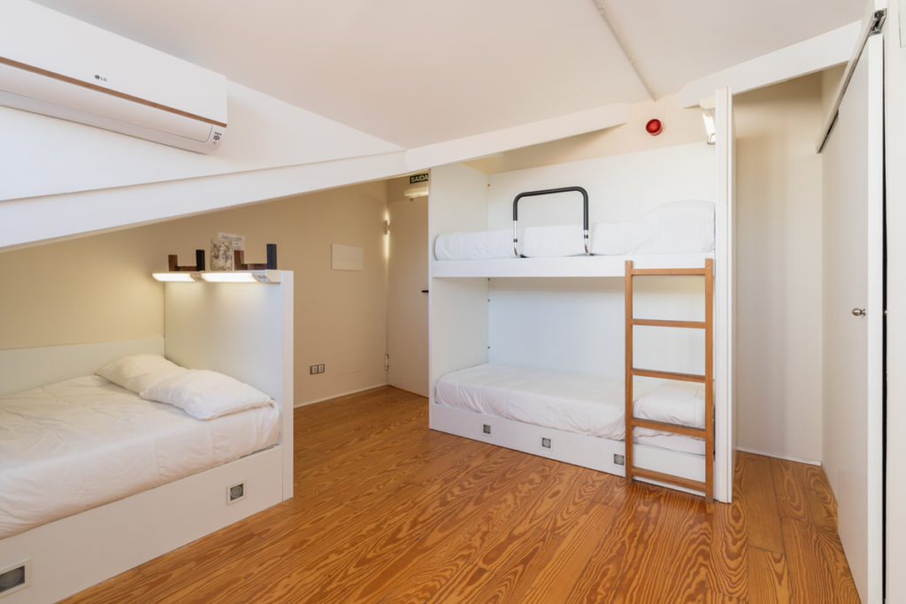Fotografía del interior de una habitación en un ático con una cama intividual y una litera con luz personalizada, y aire acondicionado.