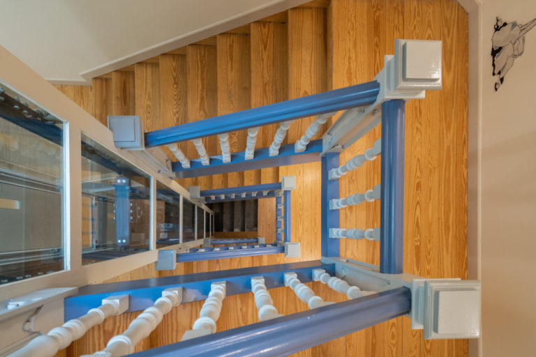 Fotografía del núcleo de escaleras de madera con pasamanos pintado de azul y ascensor acristalado.